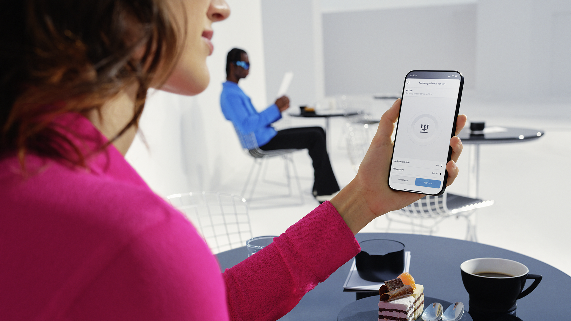 Nainen kahvilassa puhelin kädessä. Puhelimessa Mercedes me-sovelluksen esi-ilmastoinnin aktivointinäkymä.